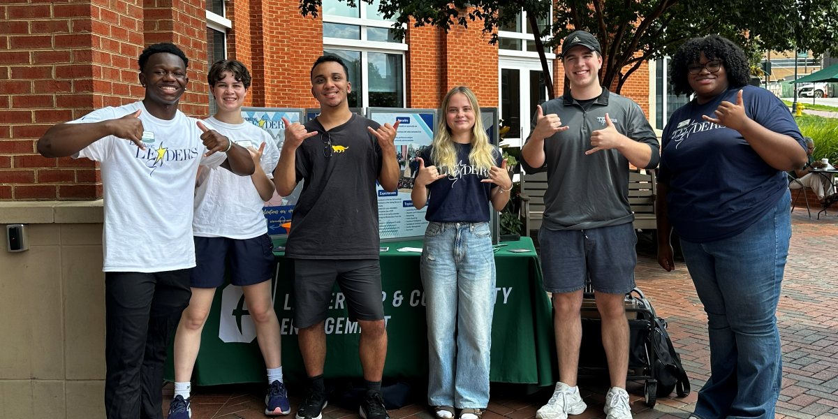 Six students, several in Emerging Leaders shirts, give shaka signs beside a Leadership and Community Engagement table outside a brick campus building