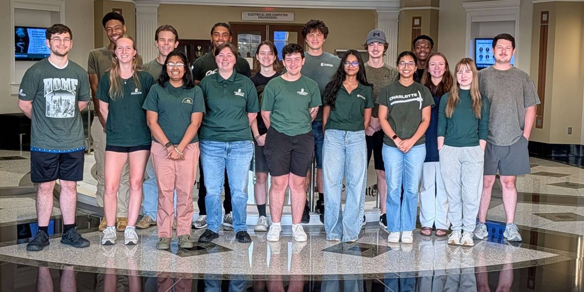 Eighteen students, most in green UNC Charlotte shirts, stand together in the lobby of an engineering building