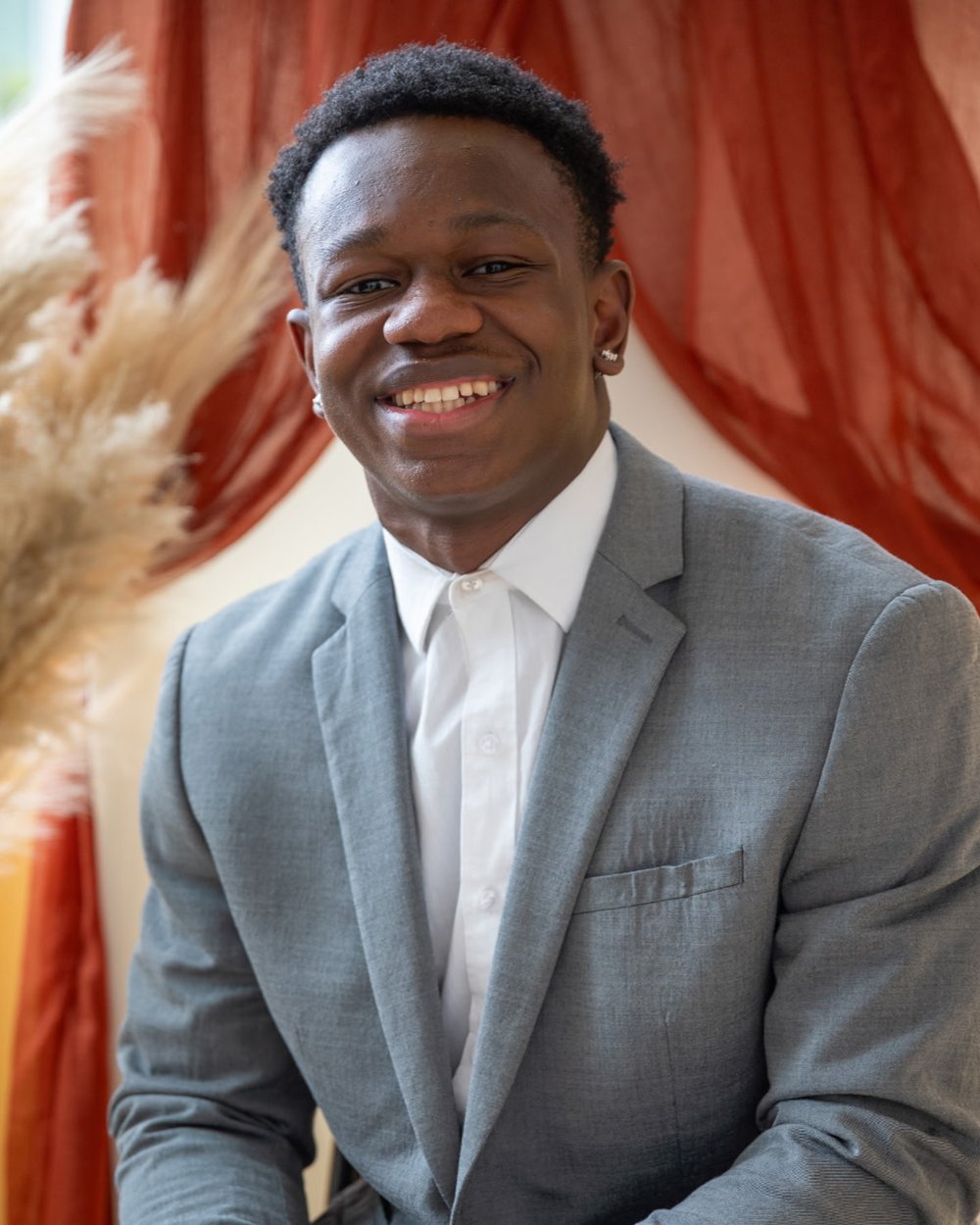 Mike Bloomfield Jr. smiling, seated in a gray blazer and white shirt in front of a rust-red curtain