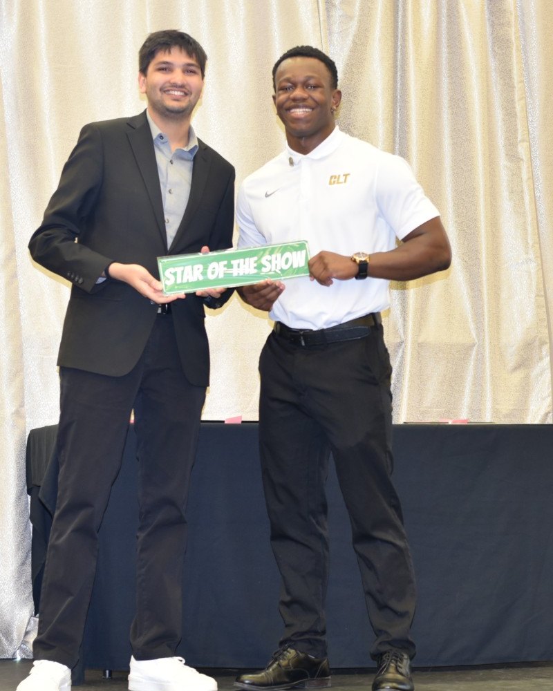 On a stage, a presenter in a black blazer and a student in a white polo smile and hold a green “Star of the Show” sign